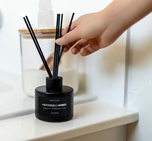 Hand placing reeds in a black diffuser bottle labeled Patchouli & Amber.
